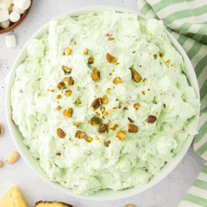 A white bowl filled with light green pistachio salad topped with chopped pistachios, surrounded by mini marshmallows, a striped green towel, and other classic pistachio salad ingredients on a light surface.