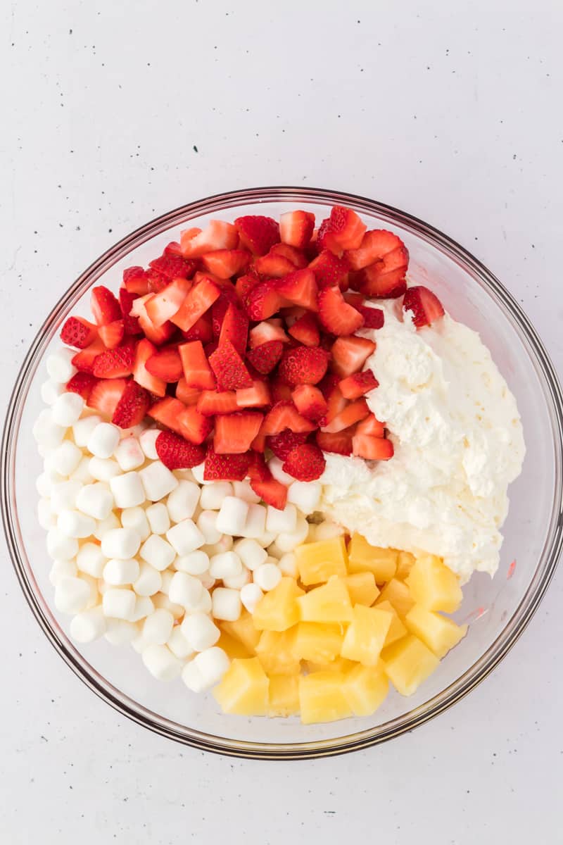 ingredients for strawberry fluff salad in a glass bowl including mini marshmallows, pineapple chunks, cool whip and chopped strawberries