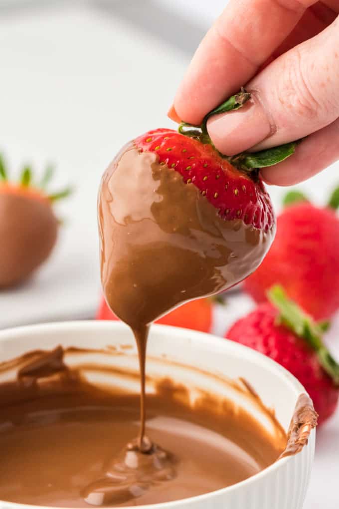 A strawberry that has been dipped in melted chocolate being held over the bowl of chocolate, letting the excess chocolate drizzle off