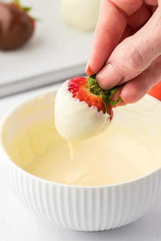 A strawberry that has been dipped in melted white chocolate being held over the bowl of chocolate, letting the excess chocolate drizzle off