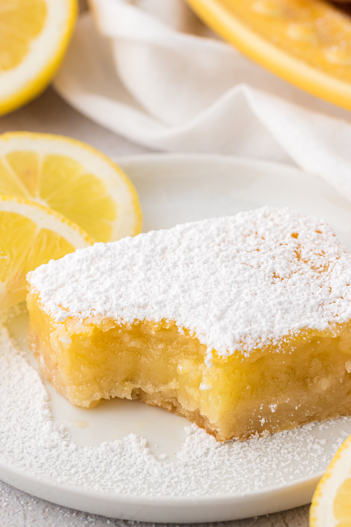 a lemon bar slice with one bite missing on a white plate dusted with powdered sugar and surrounded by lemon slices