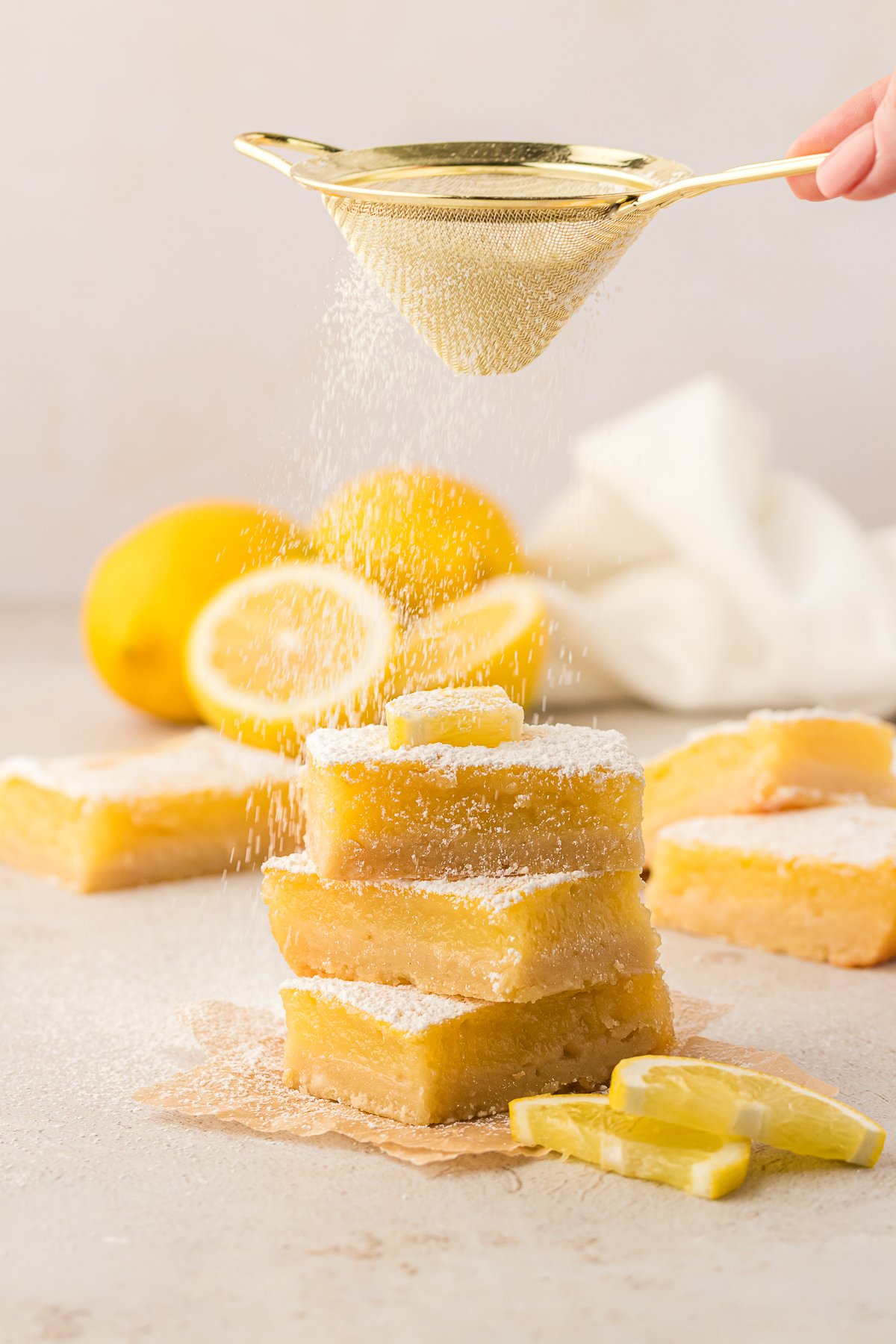 a stack of three lemon bar squares being dusted with powdered sugar surrounded by fresh lemon slices, whole lemons and more lemon bars