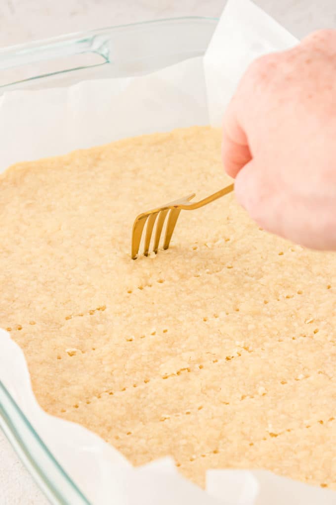 a hand poking holes with a fork into lemon bar crust inside a glass pan lined with parchment paper
