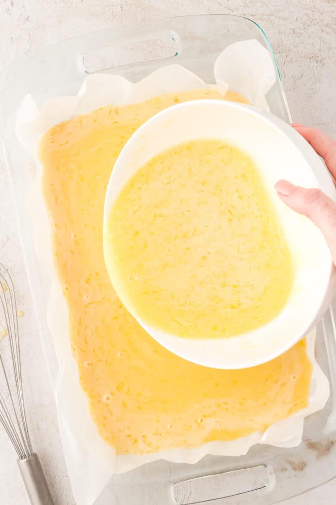 lemon filling mixture being poured over a shortbread crust in a glass baking pan lined with parchment paper
