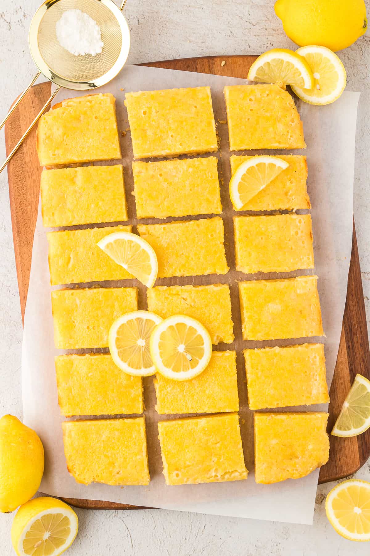sliced lemon bar squares on a wooden cutting board topped with parchment paper, surrounded by lemon slices, whole lemons, and powdered sugar