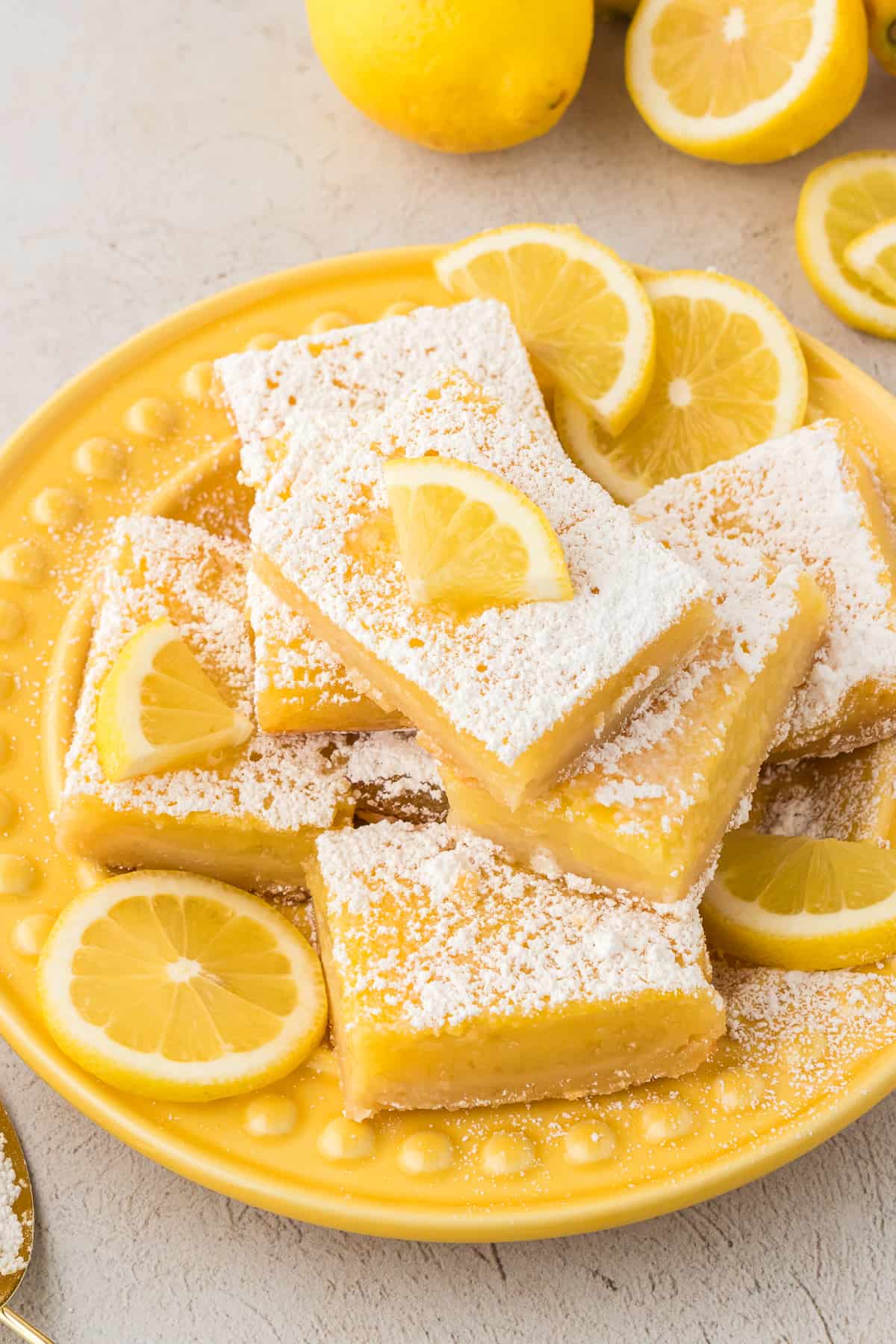 lemon bars piled on a yellow plate, dusted with powered sugar and topped with fresh lemon slices