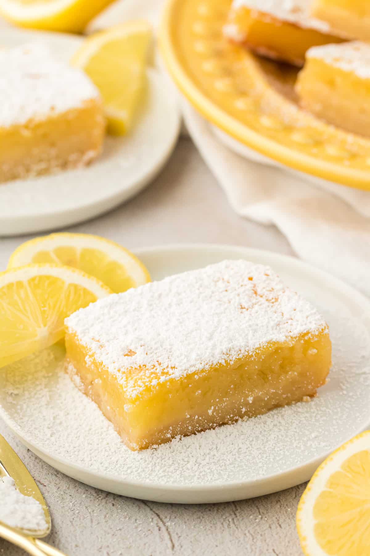 A lemon bar square on a small white plate dusted with powdered sugar and surrounded by lemon slices and whole lemons