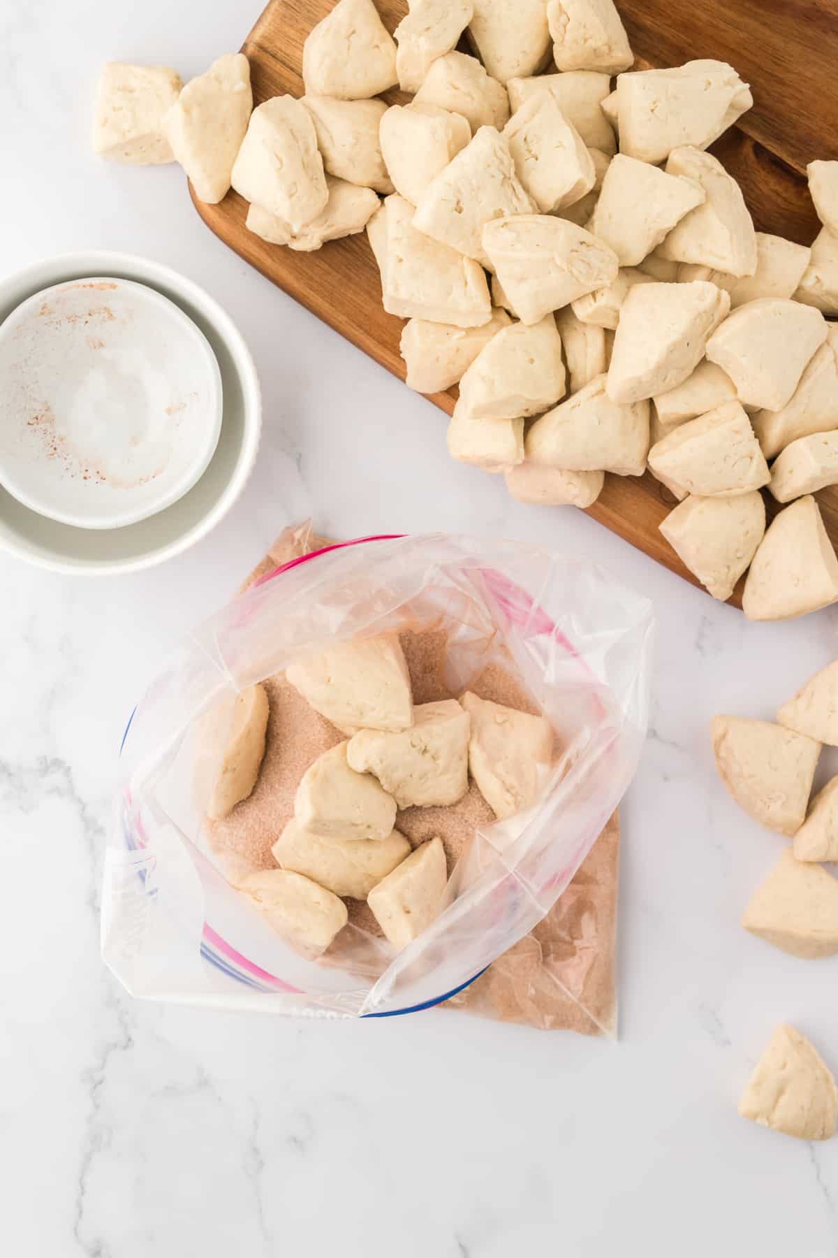 biscuit dough cut into pieces piled up on a cutting board beside a gallon plastic bag of biscuit dough pieces and with a cinnamon sugar mixture in the bottom