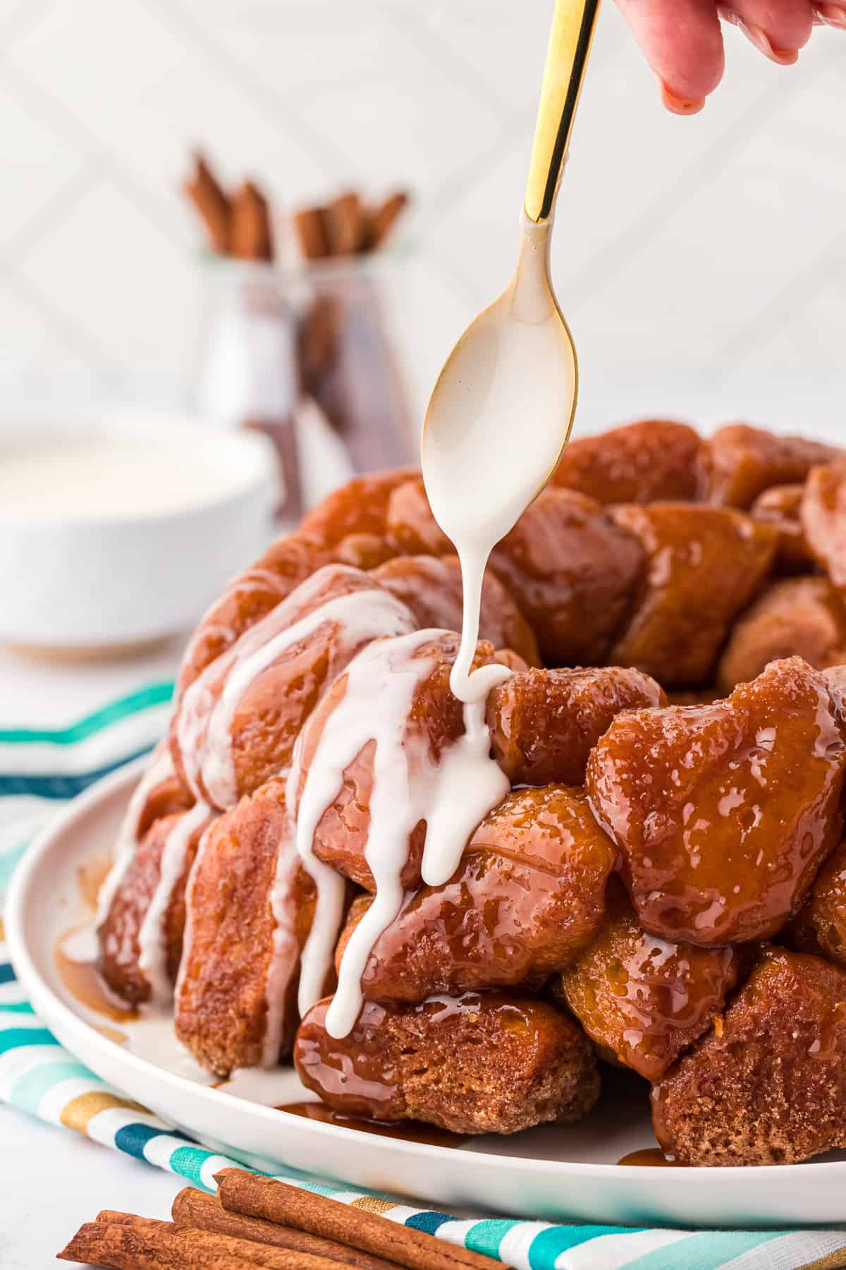 glaze being poured over a plate of monkey bread with a gold spoon, with the plate sitting on a teal, blue and gold striped towel with cinnamon sticks beside it