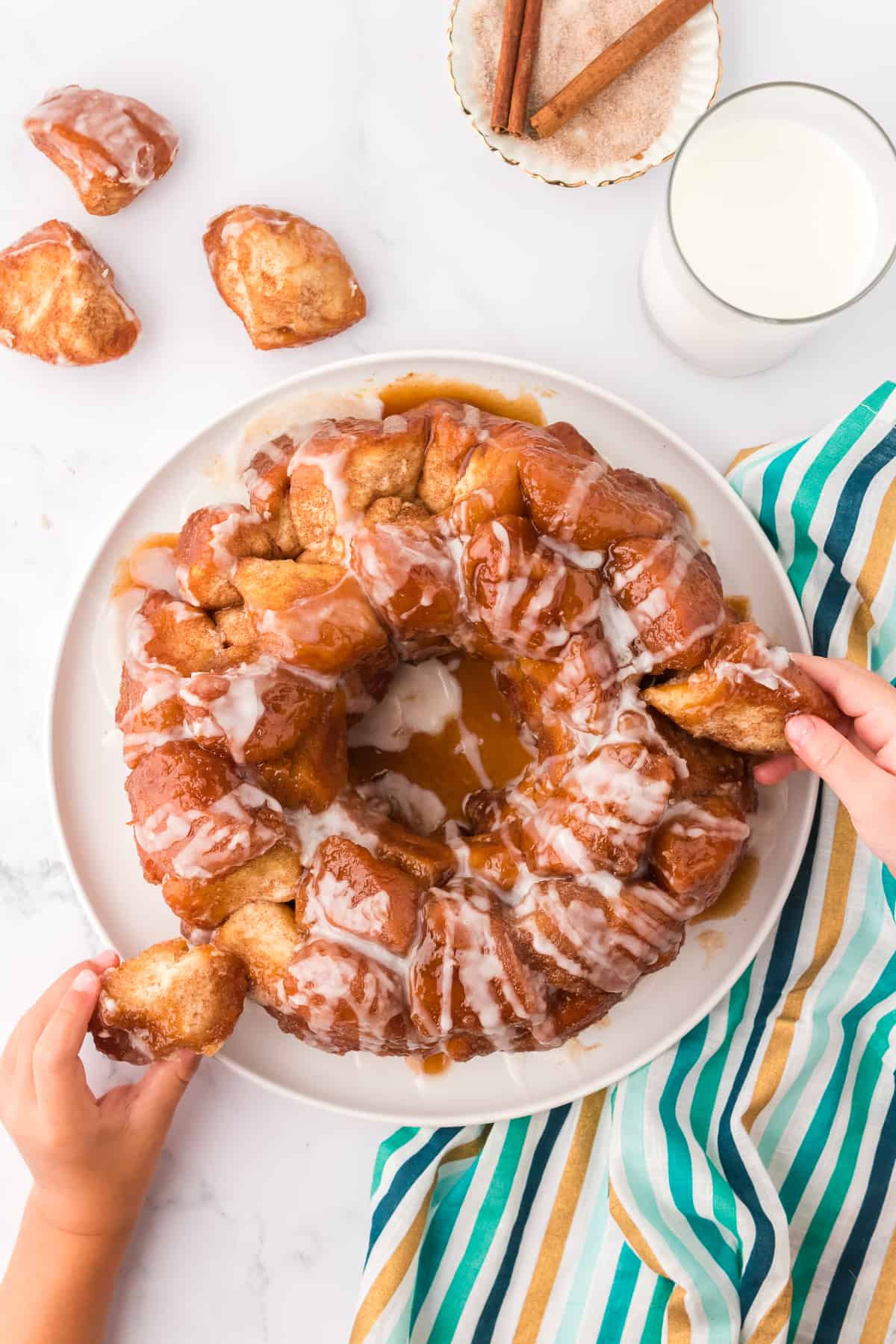 a white plate with monkey bread on it with two small hands pulling pieces off the bread, a teal, gold and blue striped towel underneath and pieces of monkey bread, milk, cinnamon sugar and cinnamon sticks arranged around it