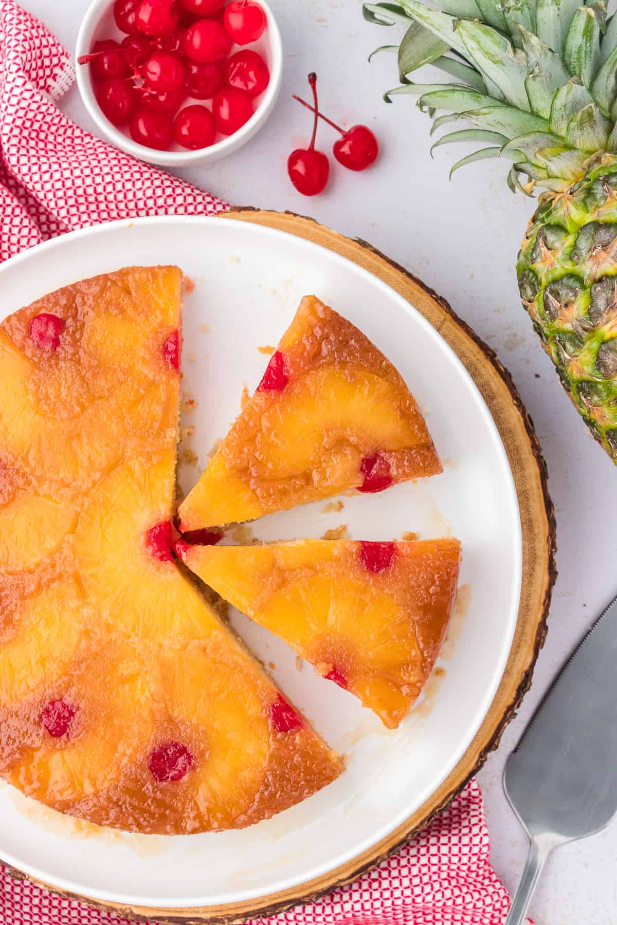 sliced pineapple upside down cake on a white plate with a red and white kitchen towel, serving spatula, pineapple and small bowl of maraschino cherries arranged around it