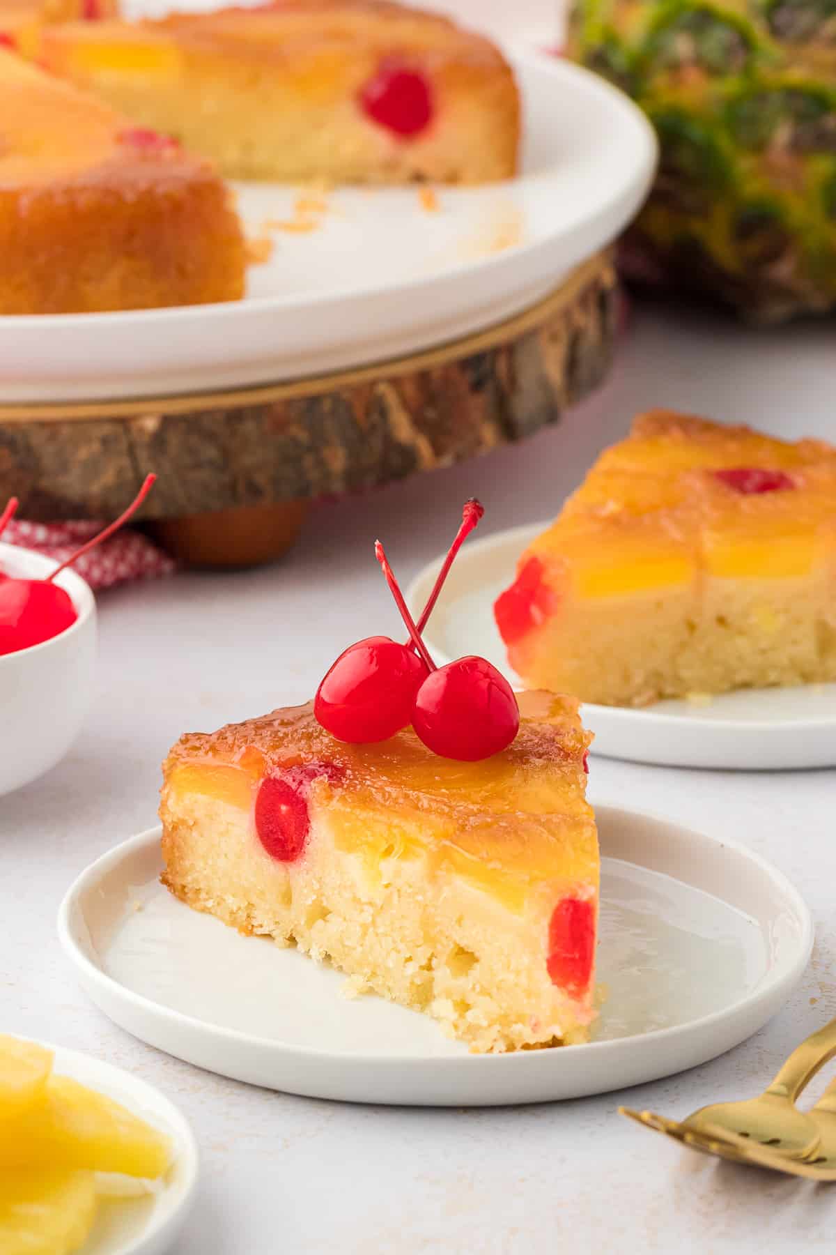 two small white plates with pineapple upside down cake slices on them arranged in front of a large plate with the rest of the cake, two gold forks, pineapple slices and a small white bowl of cherries