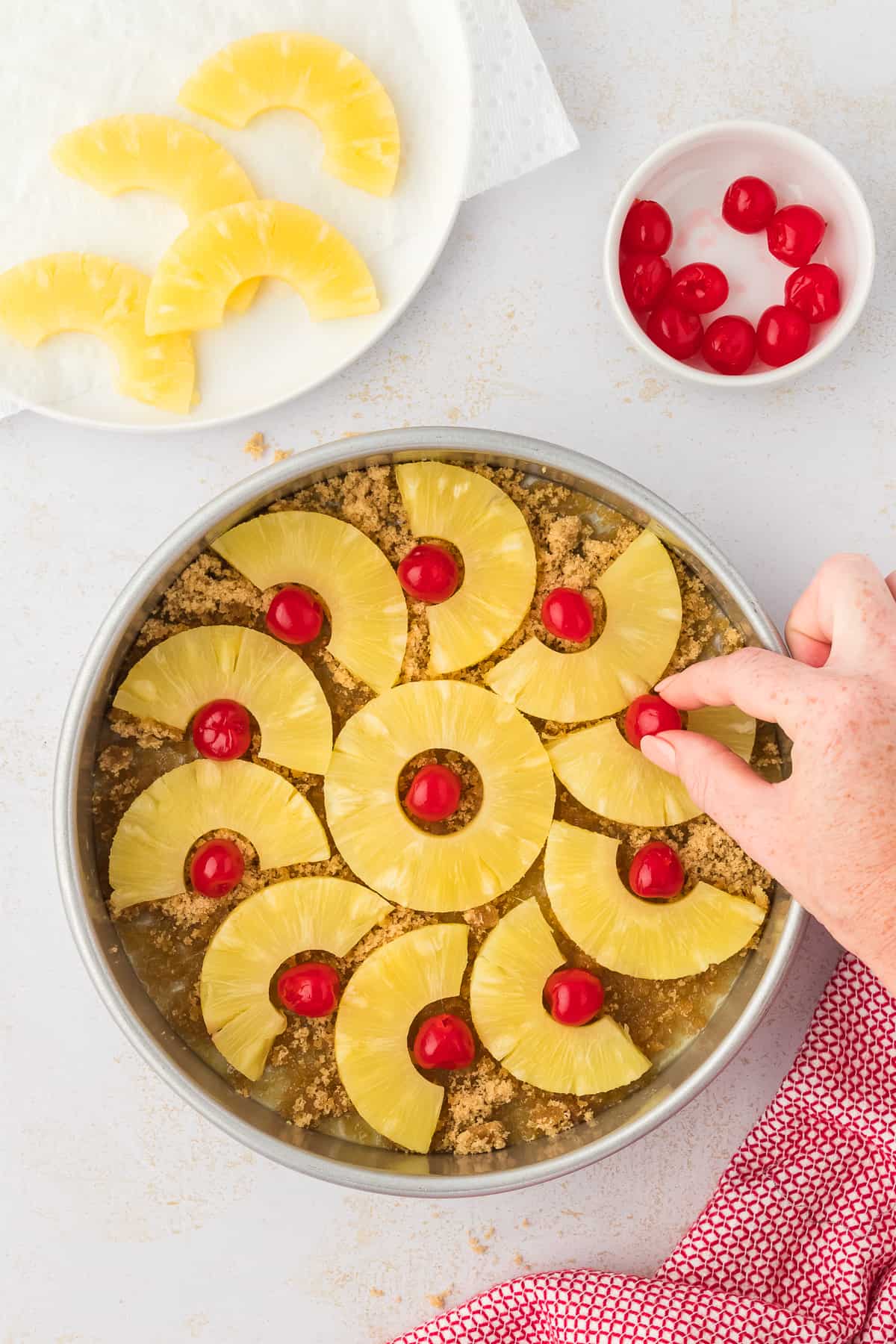 a cake pan with brown sugar and melted butter in the bottom and a hand arranging pineapple slices and maraschino cherries in it, beside a red and white kitchen towel, a plate of sliced pineapple and a small bowl of cherries