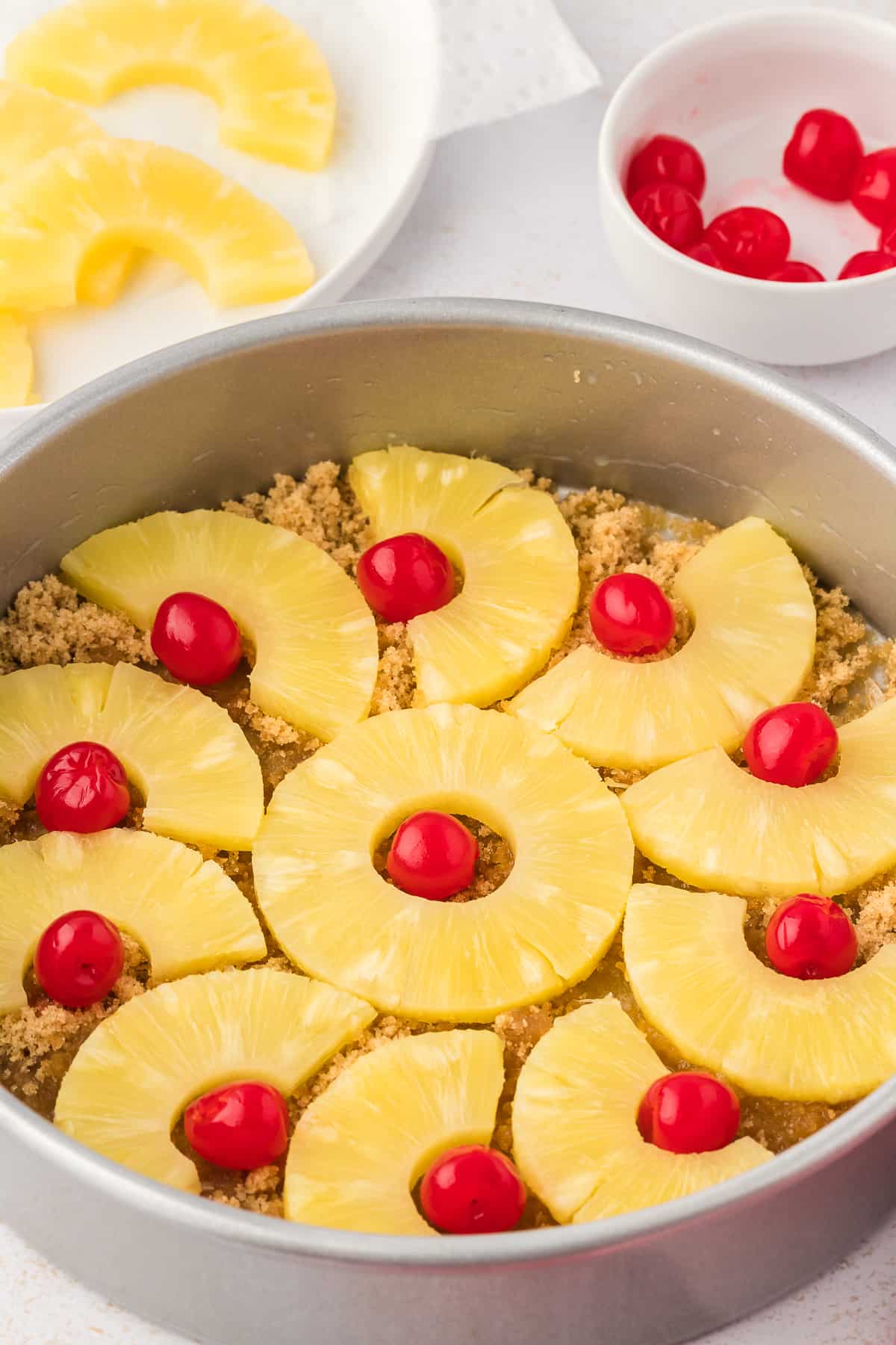 a cake pan with brown sugar and melted butter in the bottom and pineapple slices and maraschino cherries arranged in it, beside a red and white kitchen towel, a plate of sliced pineapple and a small bowl of cherries