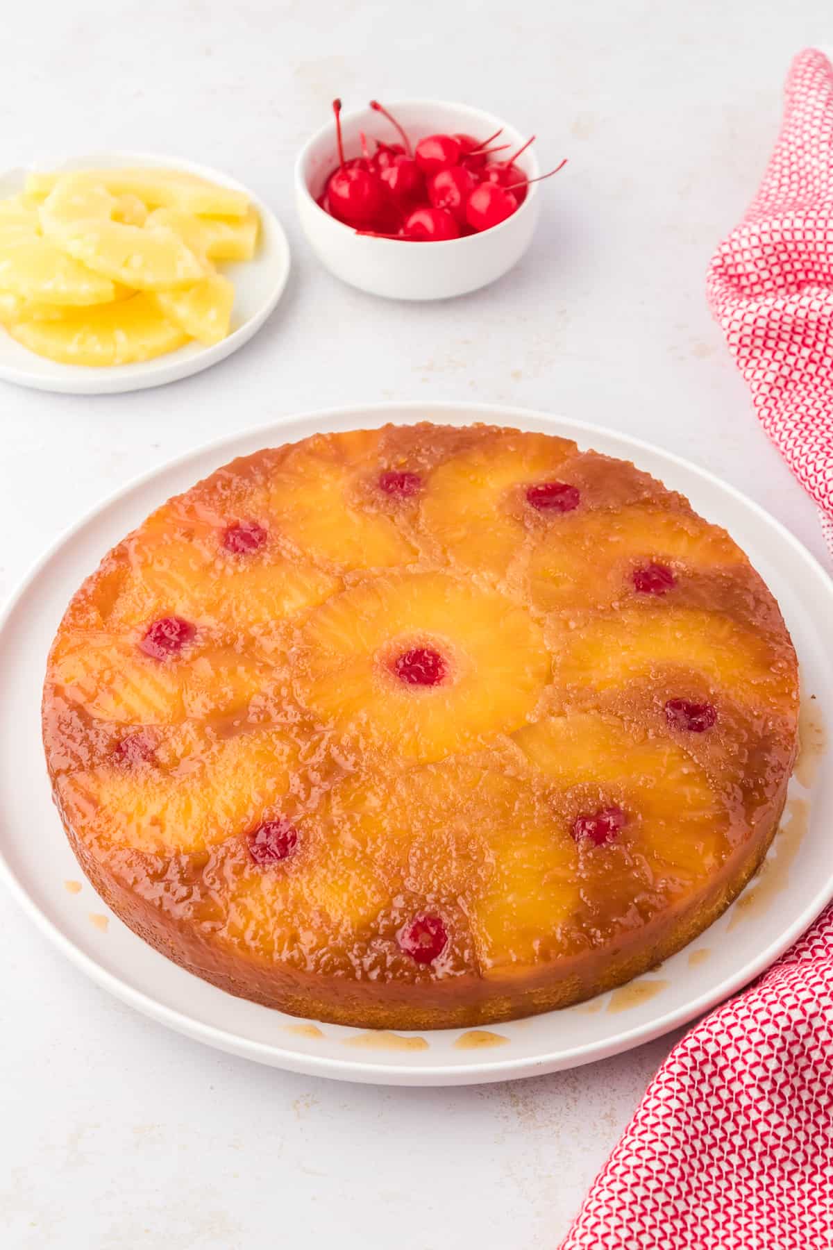 pineapple upside down cake on a white plate surrounded by pineapple slices, maraschino cherries and a red and white towel