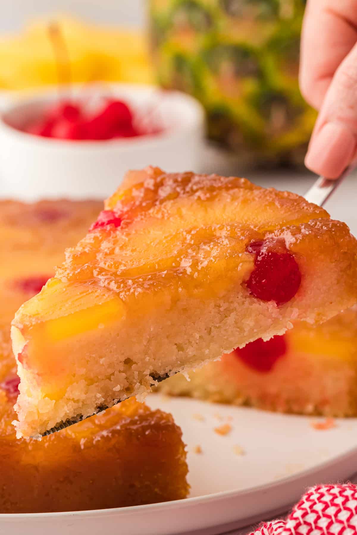 a slice of pineapple upside down cake being lifted off a plate with a pineapple, cherries and pineapple slices in the background and a red and white towel beside the plate