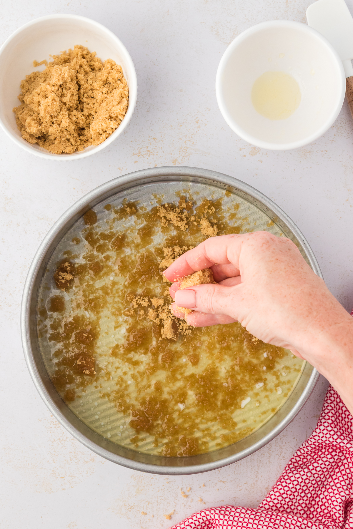 a round cake pan being sprinkled with brown sugar beside a red and white kitchen towel, a small white bowl of melted butter and another small white bowl with brown sugar in it