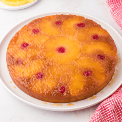 pineapple upside down cake on a white plate surrounded by pineapple slices, maraschino cherries and a red and white towel