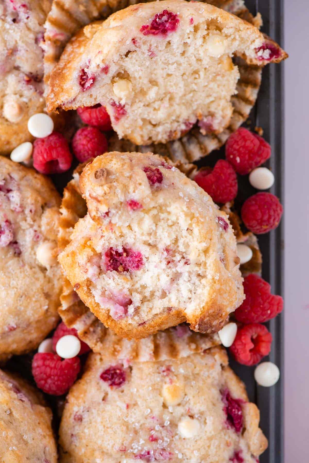 a raspberry white chocolate muffin cut in half on top of a pile of more whole raspberry muffins, fresh raspberries and white chocolate chips