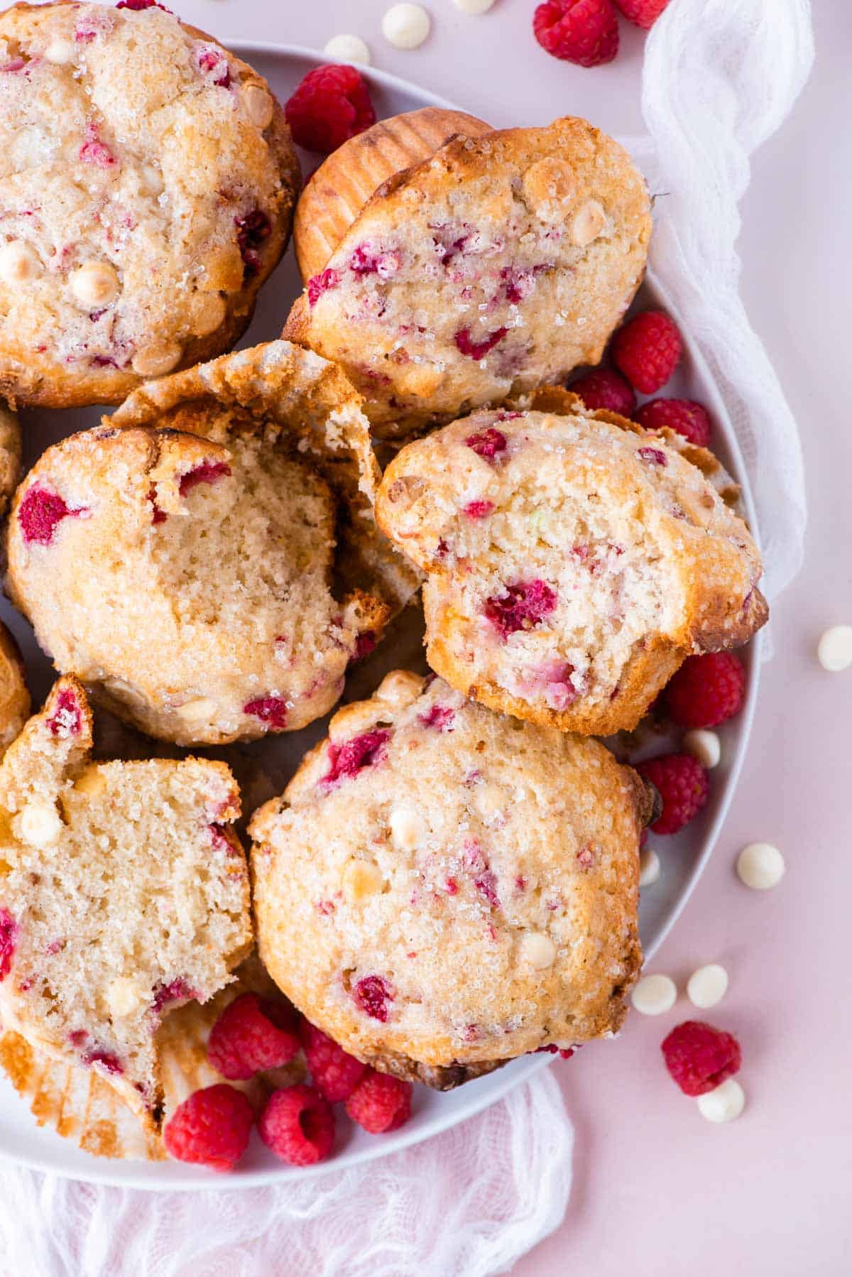 a white plate full of raspberry chocolate chip muffins on a pink surface, surrounded by fresh raspberries and white chocolate chips
