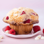 a raspberry white chocolate chip muffin on a white plate against a pink background, surrounded by fresh raspberries and white chocolate chips