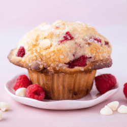 a raspberry white chocolate chip muffin on a white plate against a pink background, surrounded by fresh raspberries and white chocolate chips