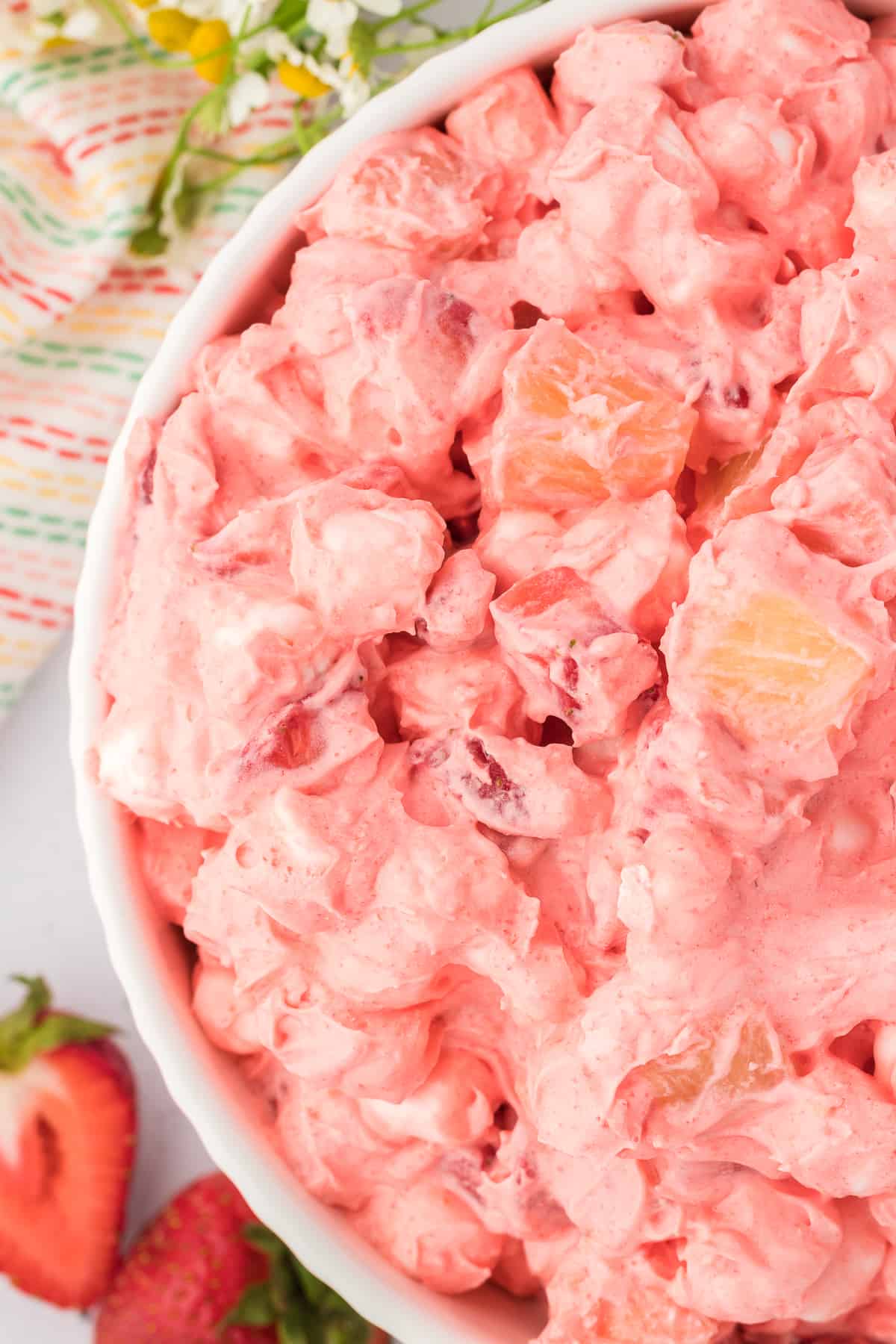 close up of the top of a bowl of strawberry fluff salad beside a colorful kitchen towel and sliced strawberries