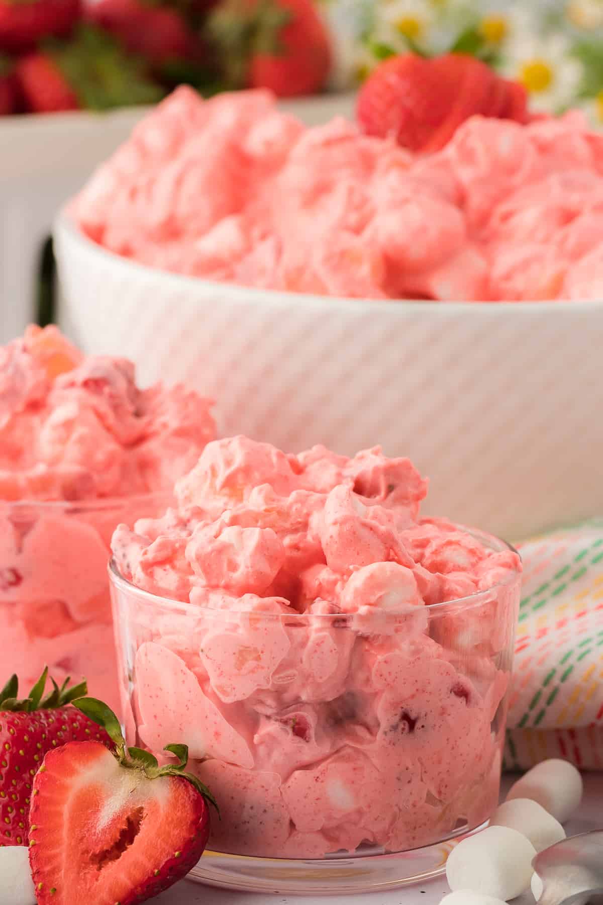 two small glass bowls of strawberry fluff surrounded by sliced strawberries, mini marshmallows, and a colorful kitchen towel, with a large white bowl of strawberry fluff in the background