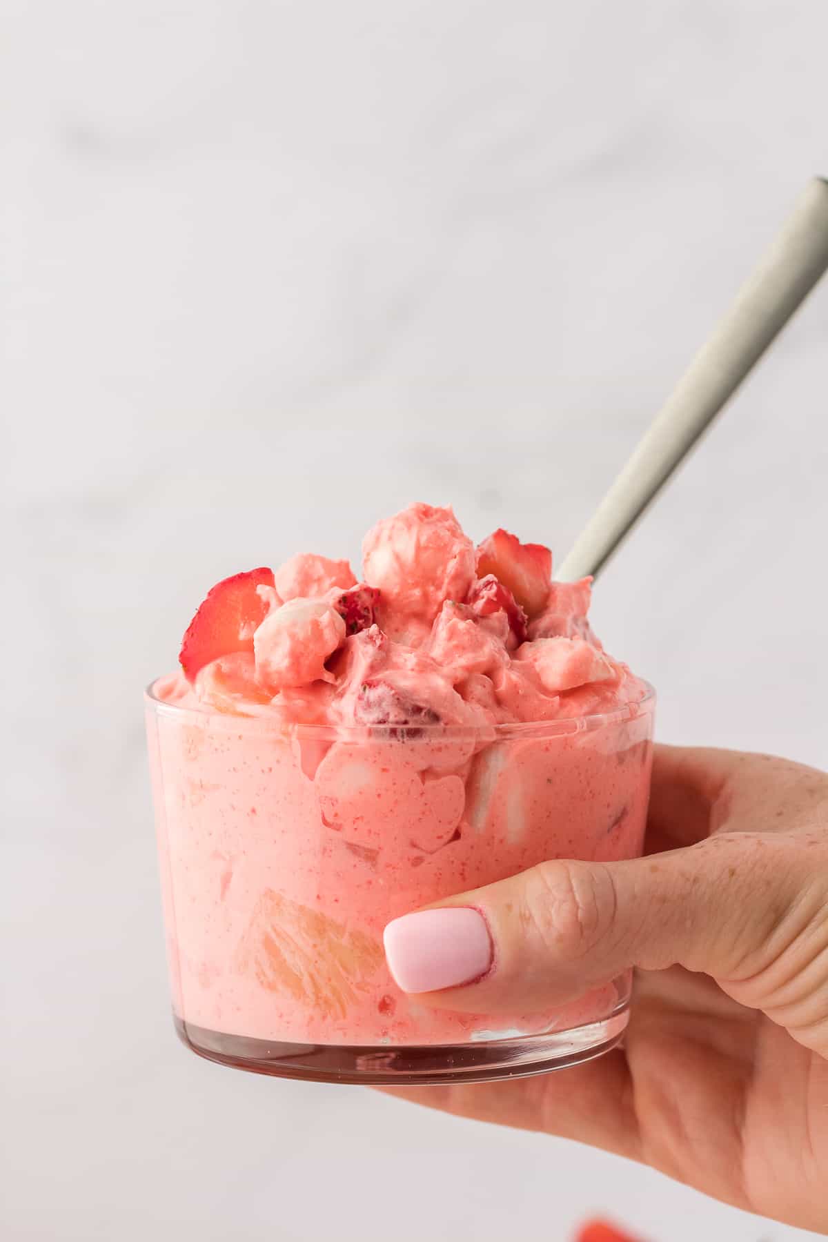 a hand holding a small glass bowl of strawberry fluff with a spoon stuck in it