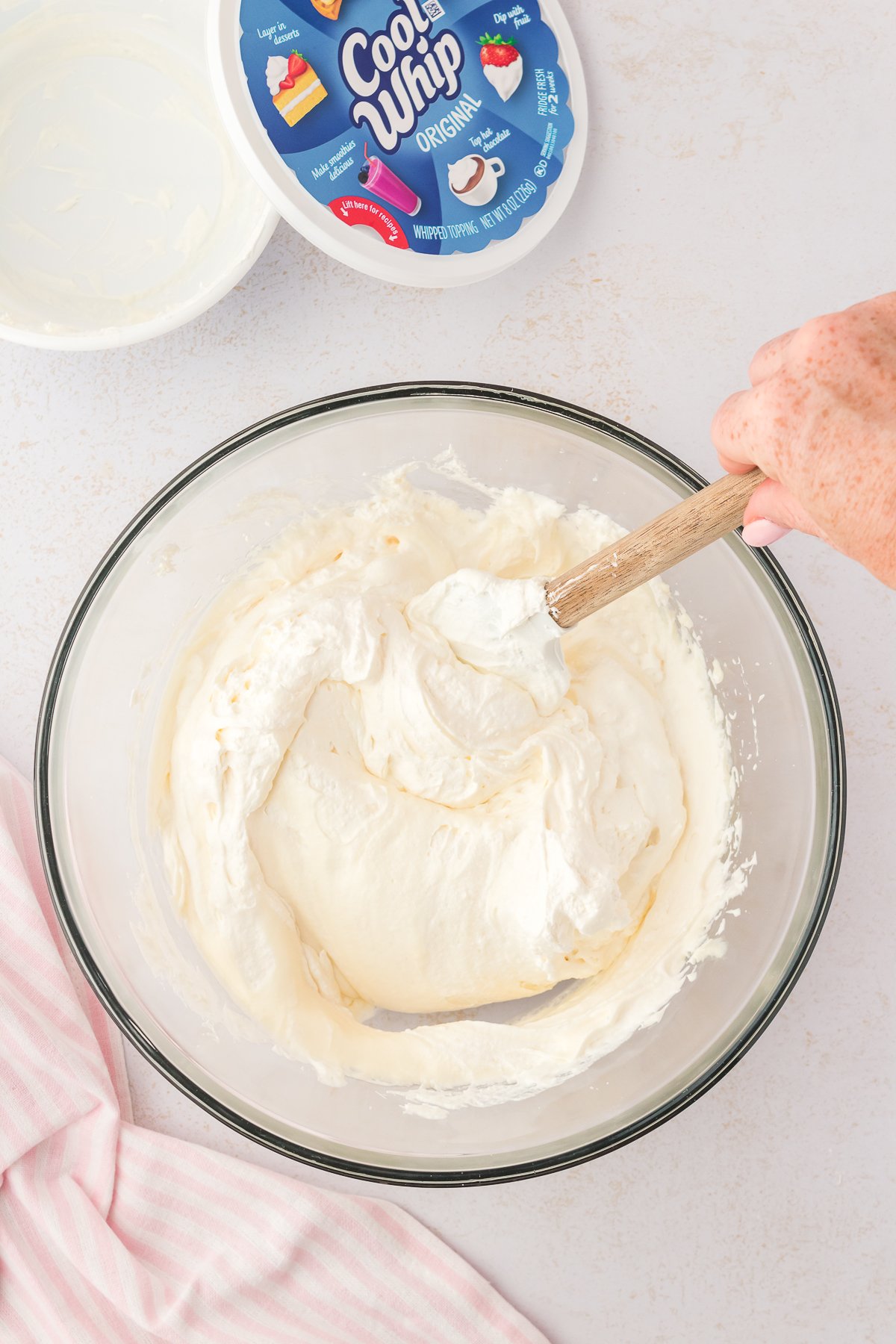 cream cheese mixture in a glass bowl being mixed with a spatula, beside a pink and white striped kitchen towel and an open container of cool whip