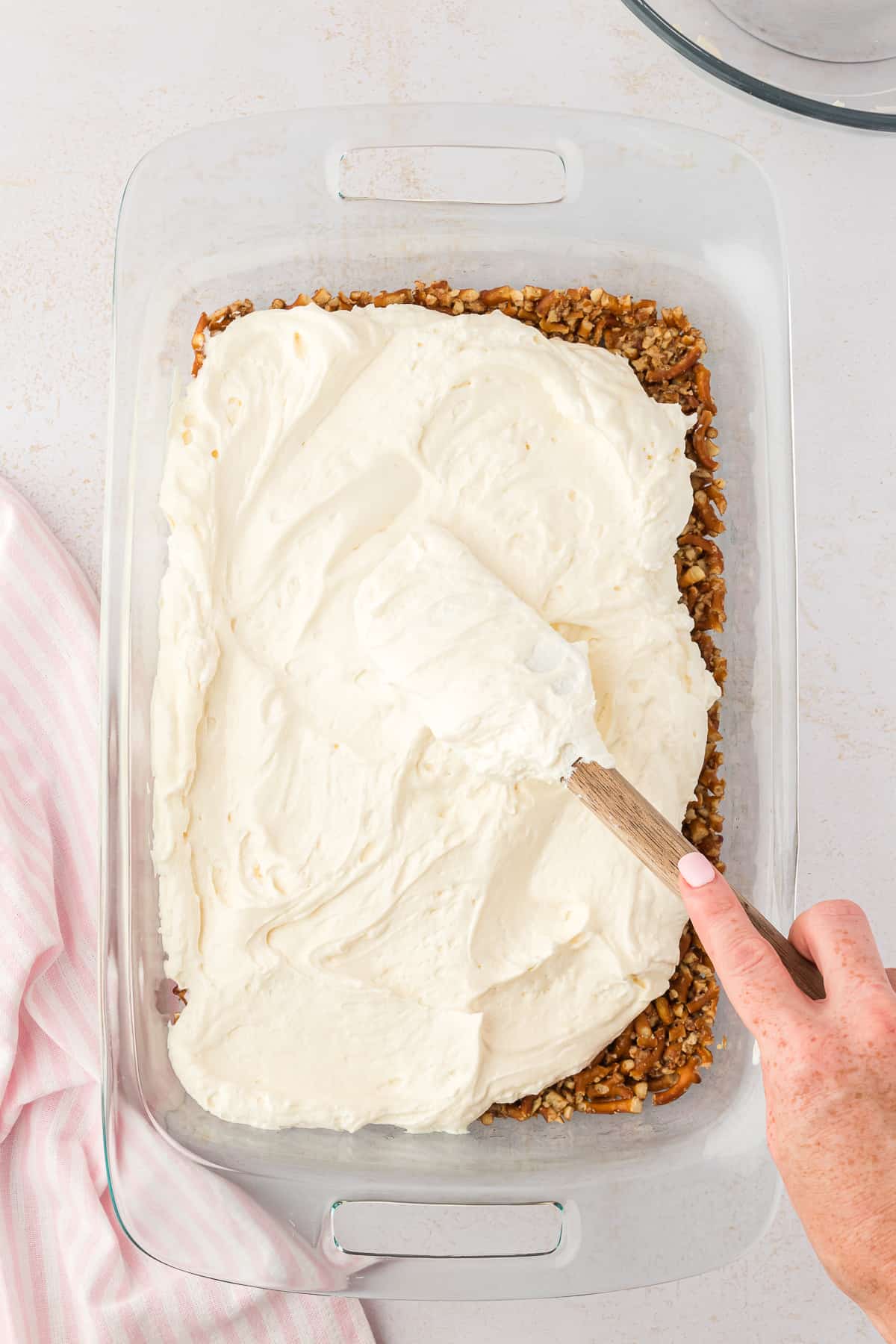the cream cheese layer of strawberry pretzel salad being spread over top of the pretzel crust layer in a large glass baking dish, beside a pink and white kitchen towel