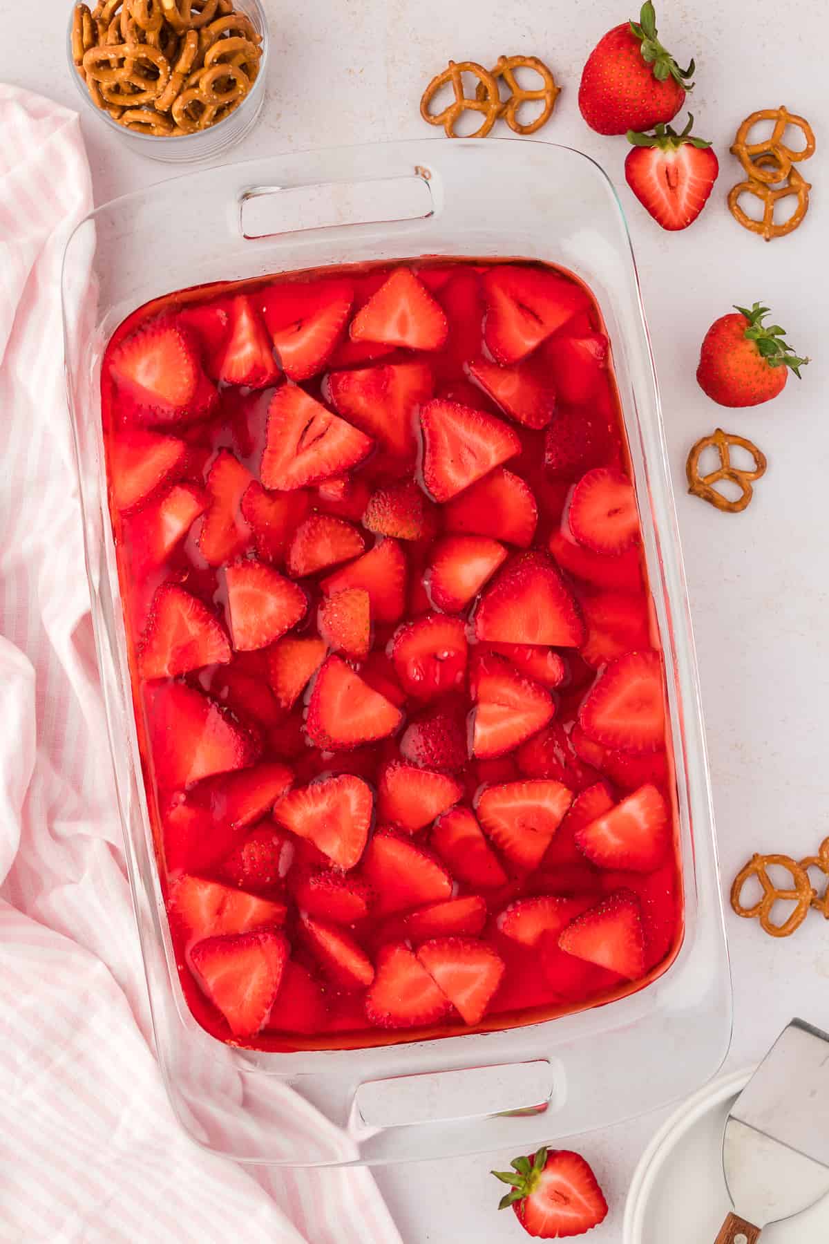 a clear glass baking dish of strawberry pretzel salad surrounded by fresh strawberries, pretzels, and a pink and white striped kitchen towel