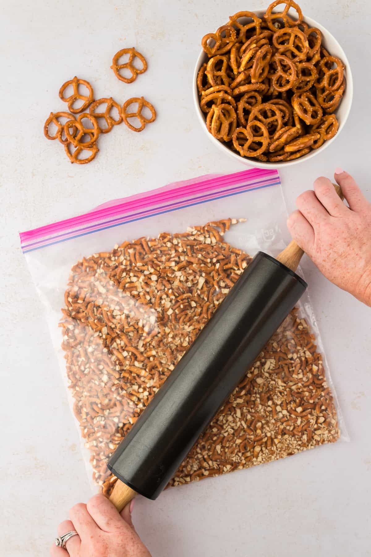 crushing pretzels in a ziploc bag with a rolling pin, with a bowl of pretzels beside is