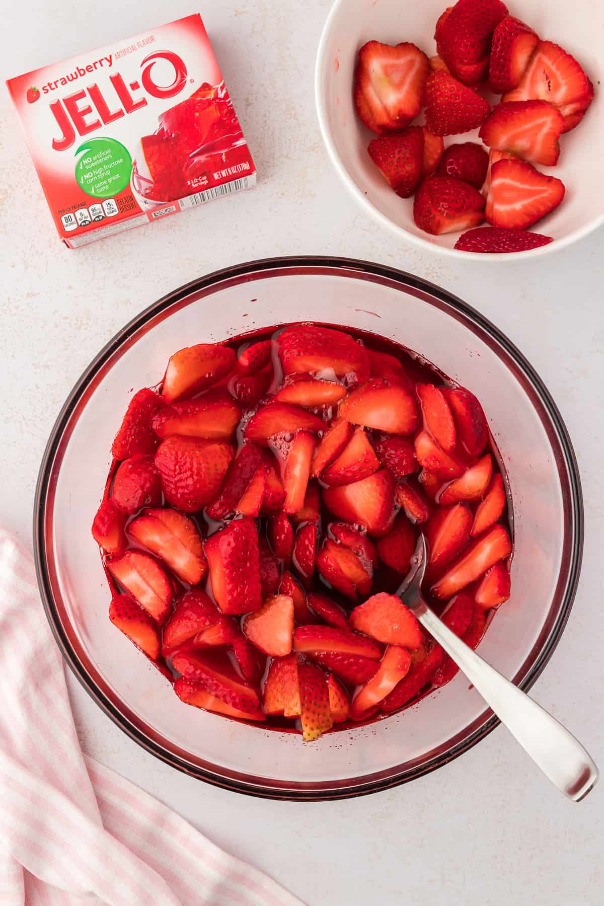 a glass bowl with the strawberry jello mixture in it for strawberry pretzel salad with a spoon in it, beside a small white bowl of fresh strawberries, a pink and white striped kitchen towel and a box of strawberry jello