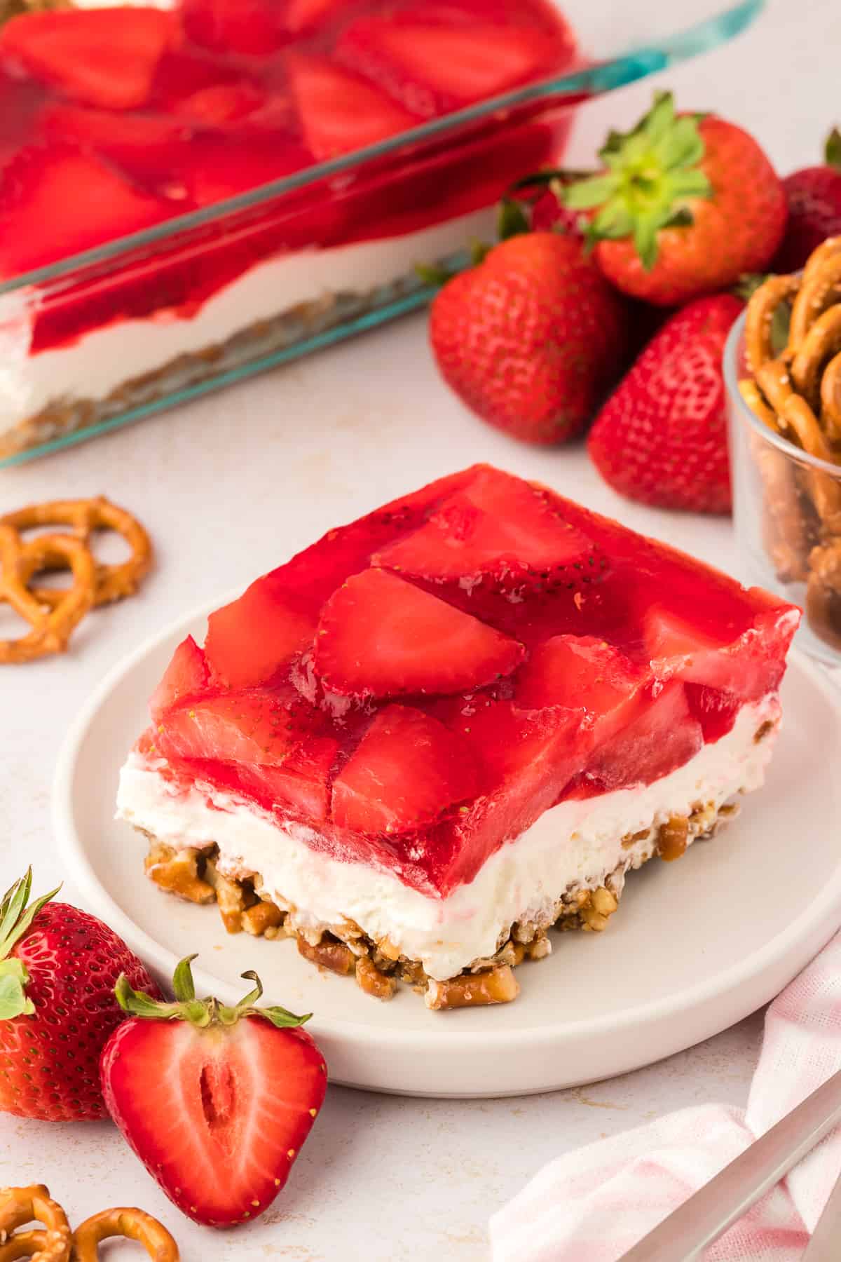 a slice of strawberry pretzel salad on a white plate, with a baking dish full of salad in the background, surrounded by fresh strawberries, pretzels and a pink and white striped kitchen towel