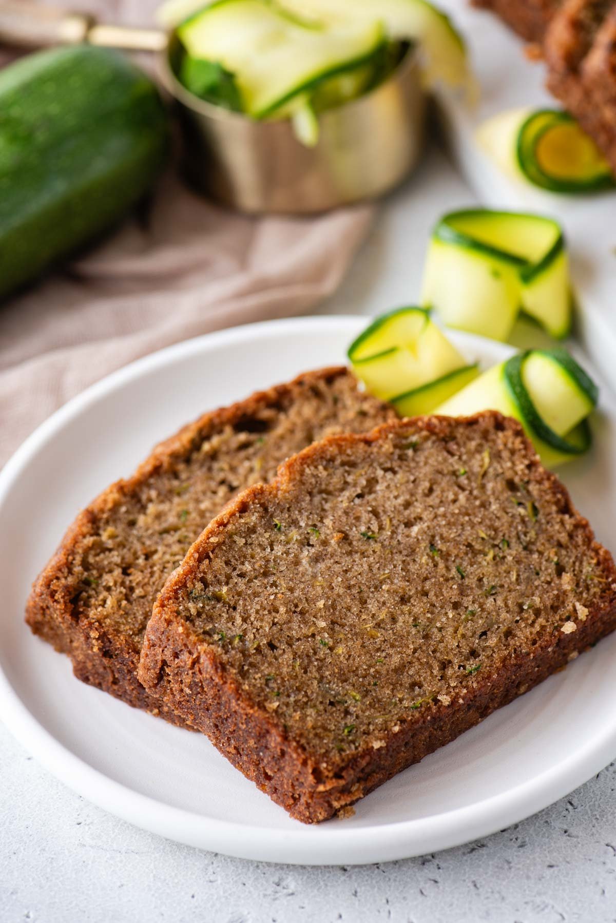 two slices of zucchini bread on a white plate with slices of fresh zucchini and whole zucchini around it