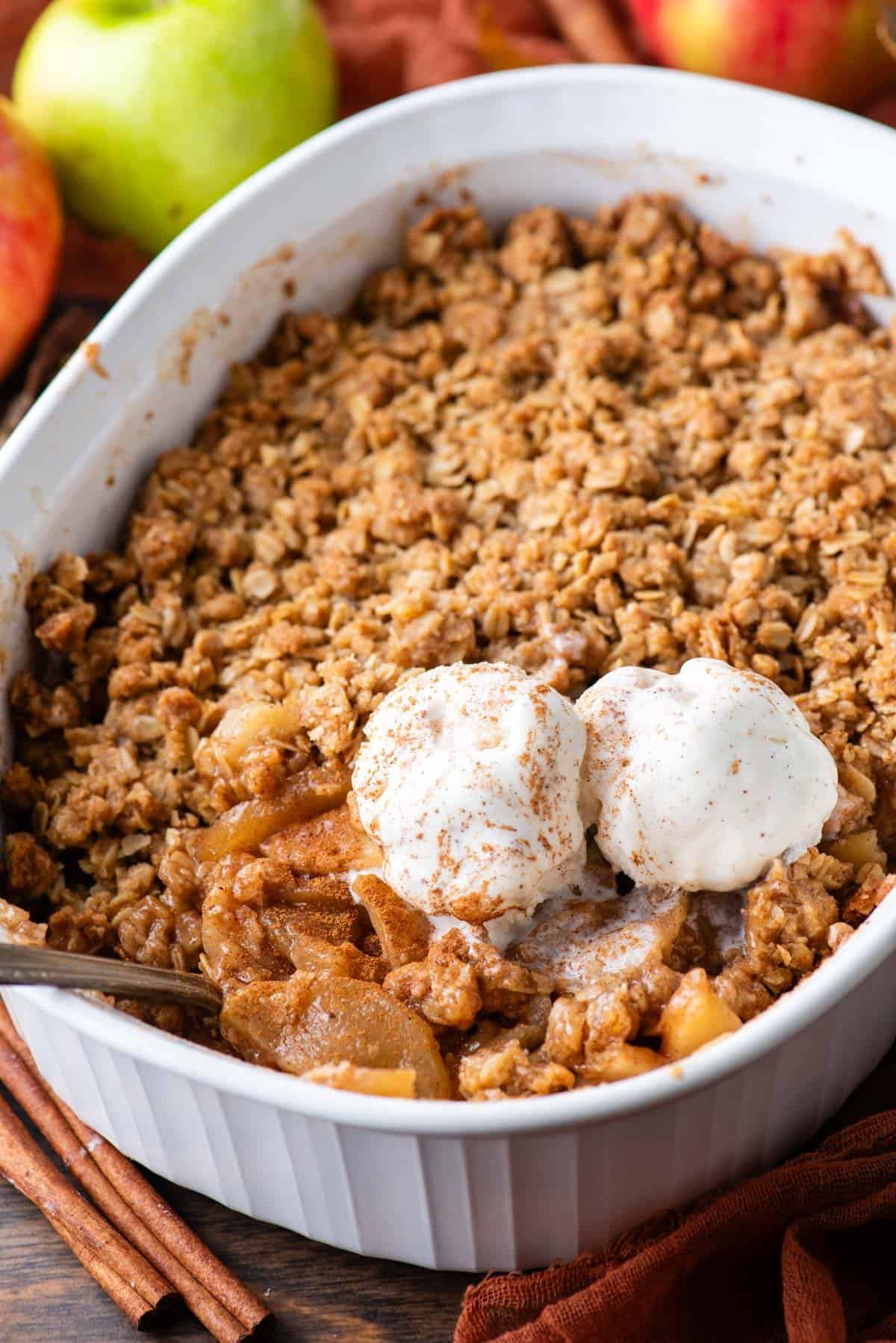 a white baking pan full of freshly baked apple crisp with a spoon in it and two scoops of vanilla ice cream
