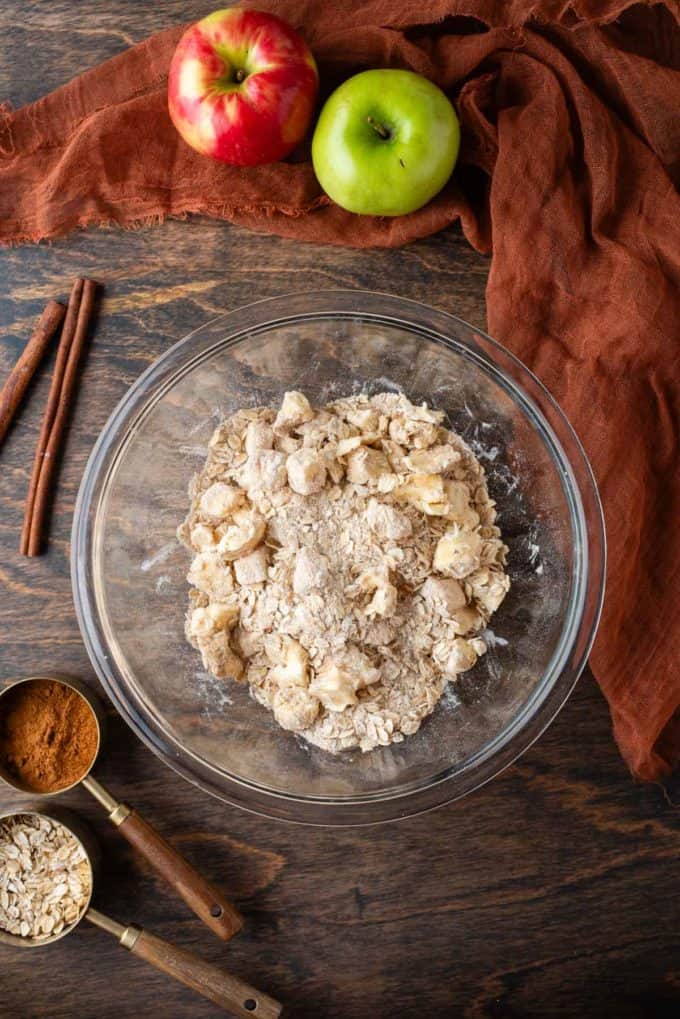 ingredients for apple crisp crumble topping in a clear glass bowl on a wood surface, surrounded by a spoon of oats, a spoon of cinnamon, cinnamon sticks, a dark orange towel and whole green apple