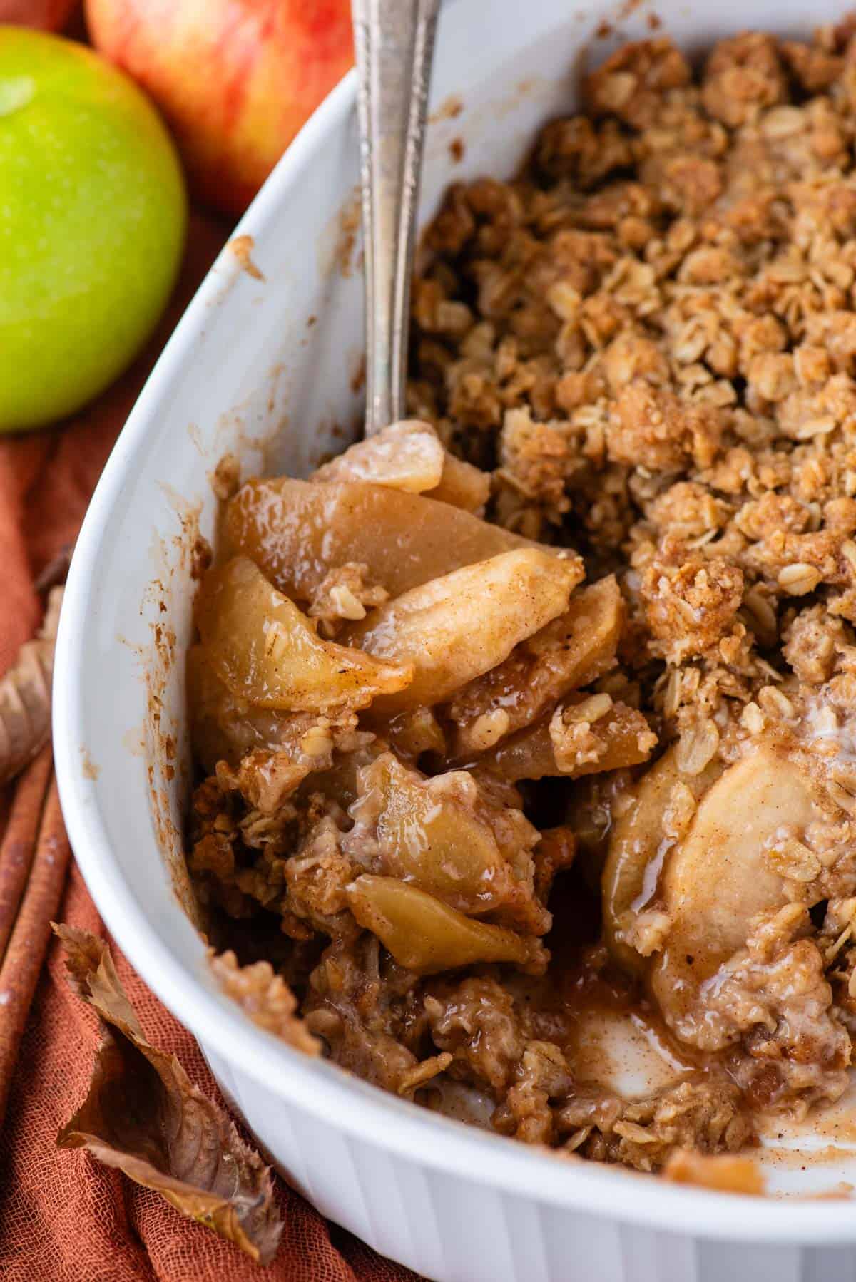 close up of a white baking pan full of apple crisp with a spoon in it and some of the crisp missing with a dark orange towel and whole green apple beside the dish