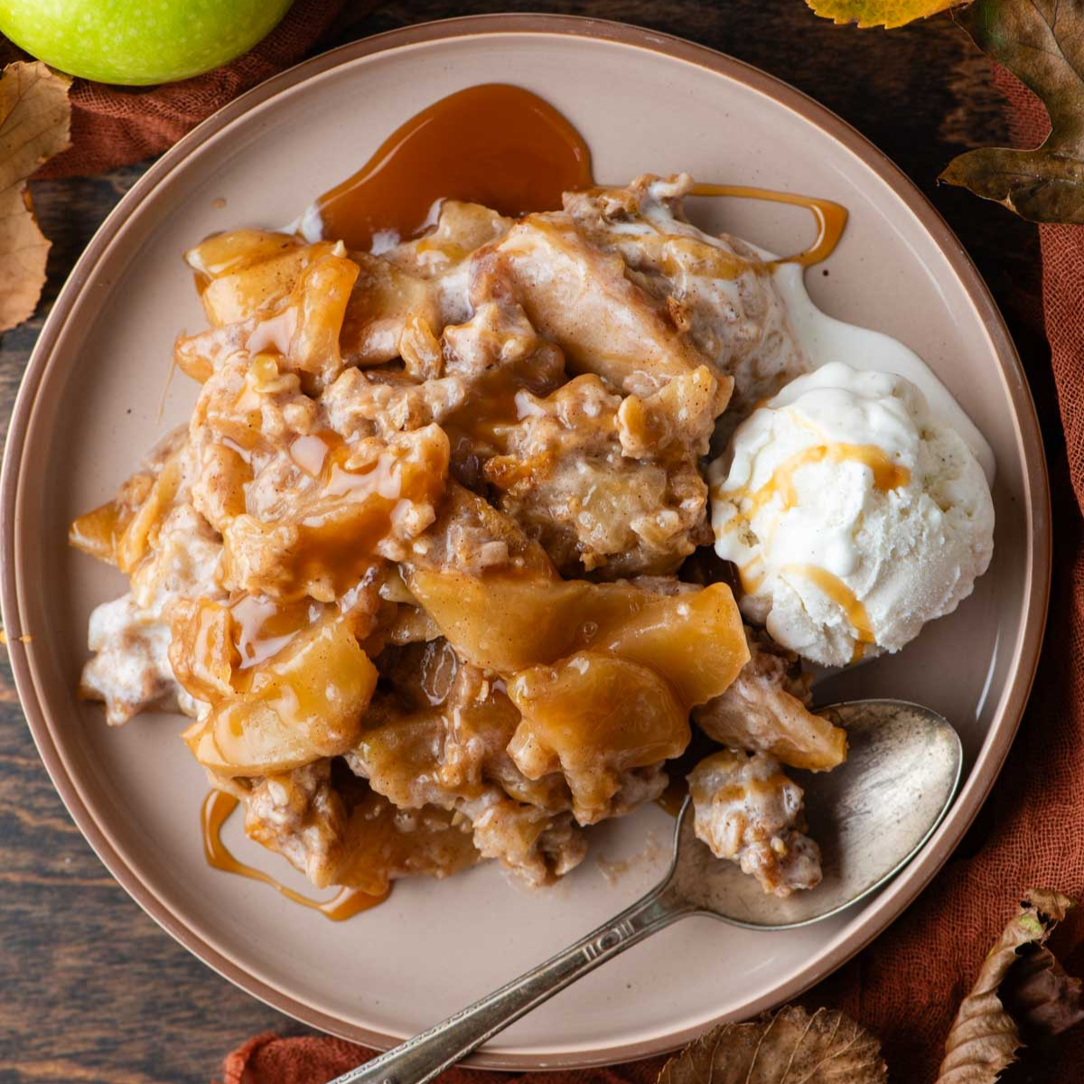 apple crisp on a plate drizzled with caramel sauce with a scoop of ice cream and a spoon beside it and fresh apples, fall leaves and an orange towel around it