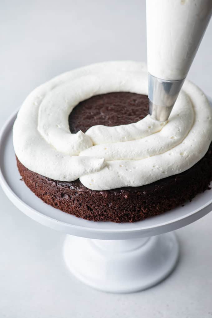 one layer of chocolate cake on a white cake stand with stabilized whipped cream being piped on top