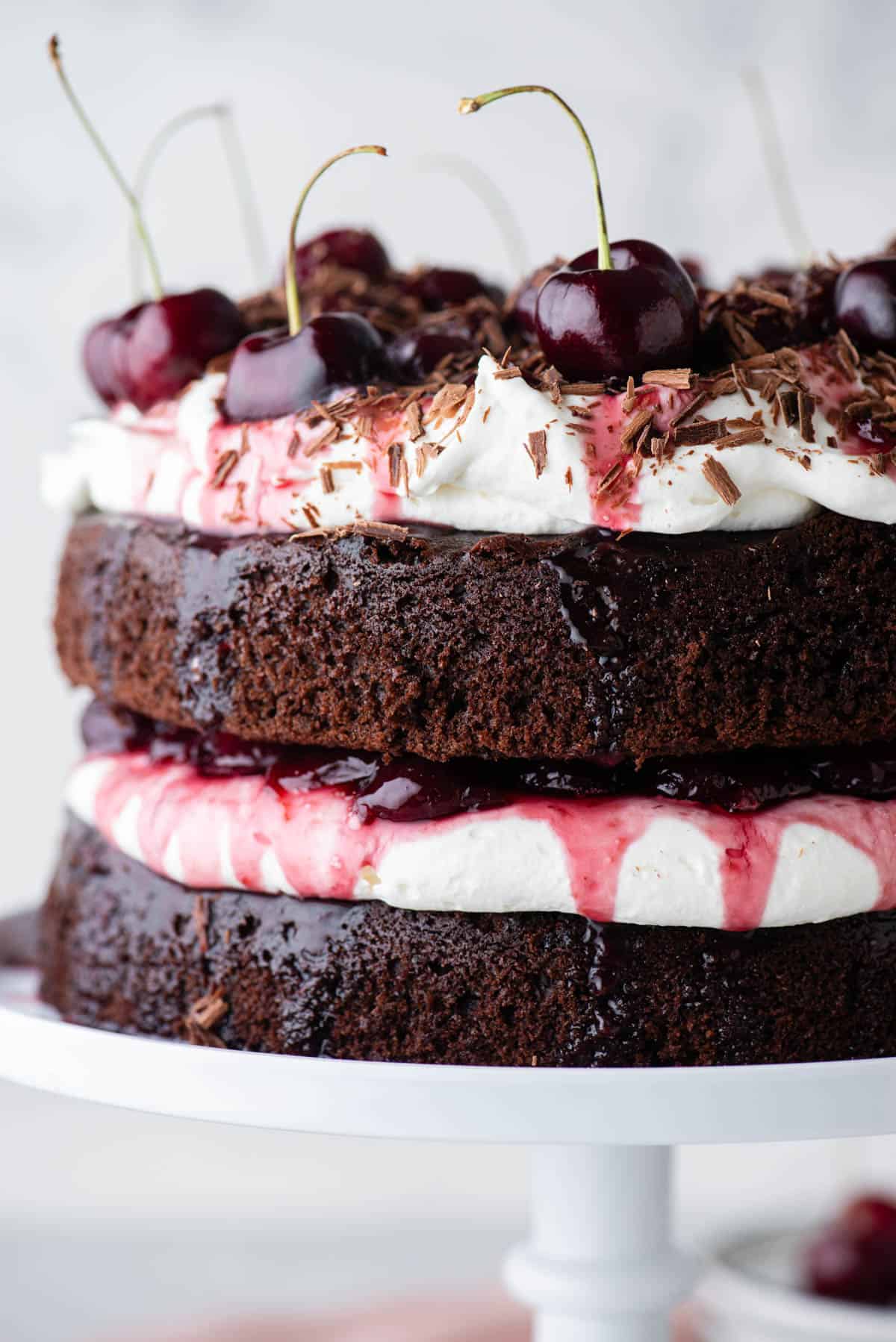 close up of the side of a black forest cake on a white cake stand with layers of chocolate cake, whipped cream, and cherry filling