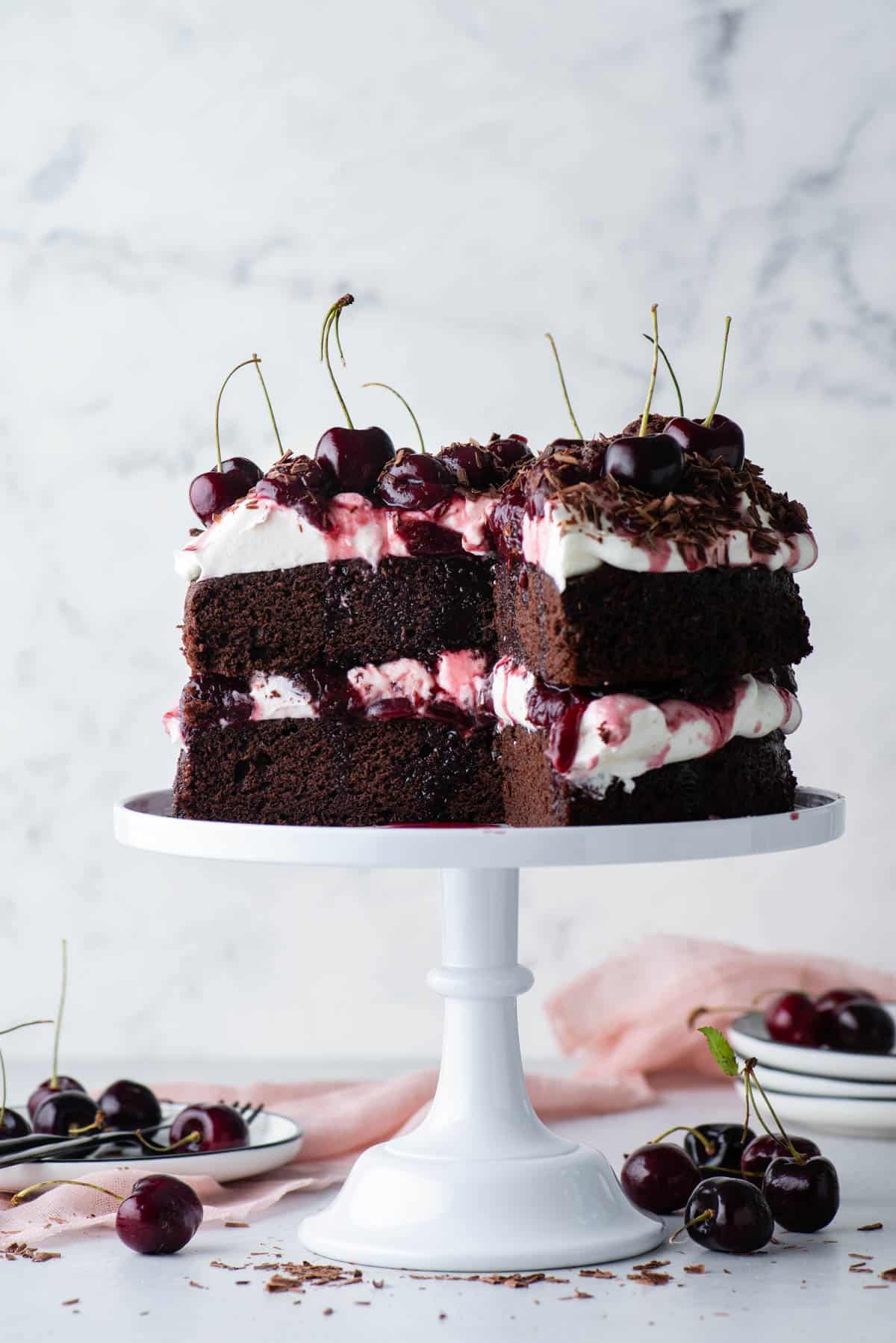 black forest cake on a white cake stand with a large slice taken out, with stacks of white plates and cherries scattered around