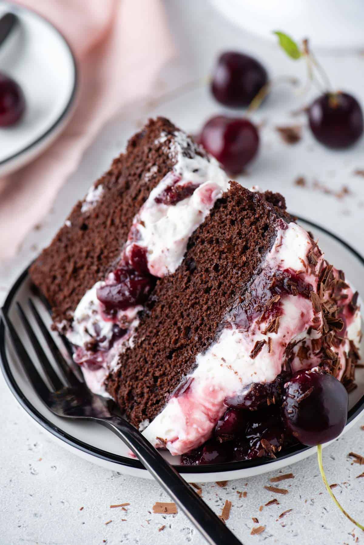 a slice of black forest ake on a plate with a fork surrounded by cherries and little pieces of shaved chocolate