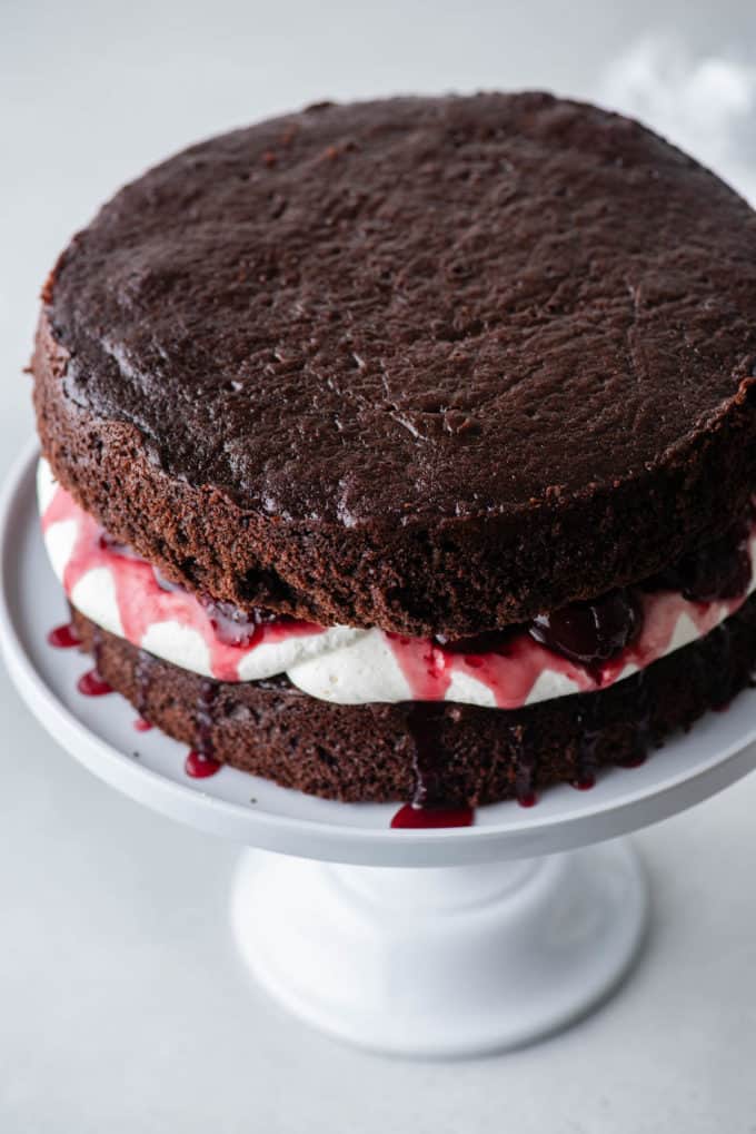one layer of chocolate cake on a white cake stand with stabilized whipped cream on top and a cherry filling on top of it, with another layer of chocolate cake on top of that