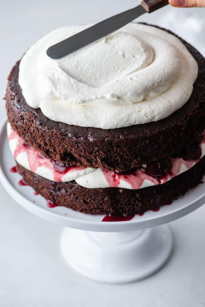 black forest cake on a white cake stand with a layer of chocolate cake, whipped cream, cherries, more chocolate cake and whipped cream being spread on top
