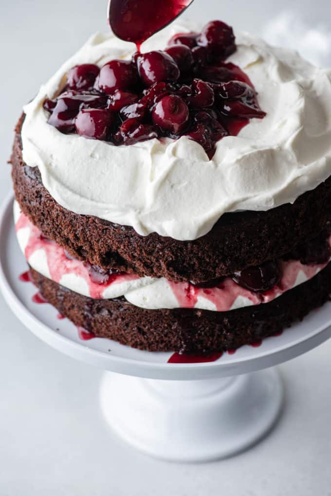 black forest cake on a white cake stand with a layer of chocolate cake, whipped cream, cherries, more chocolate cake, more whipped cream and cherries being added on top
