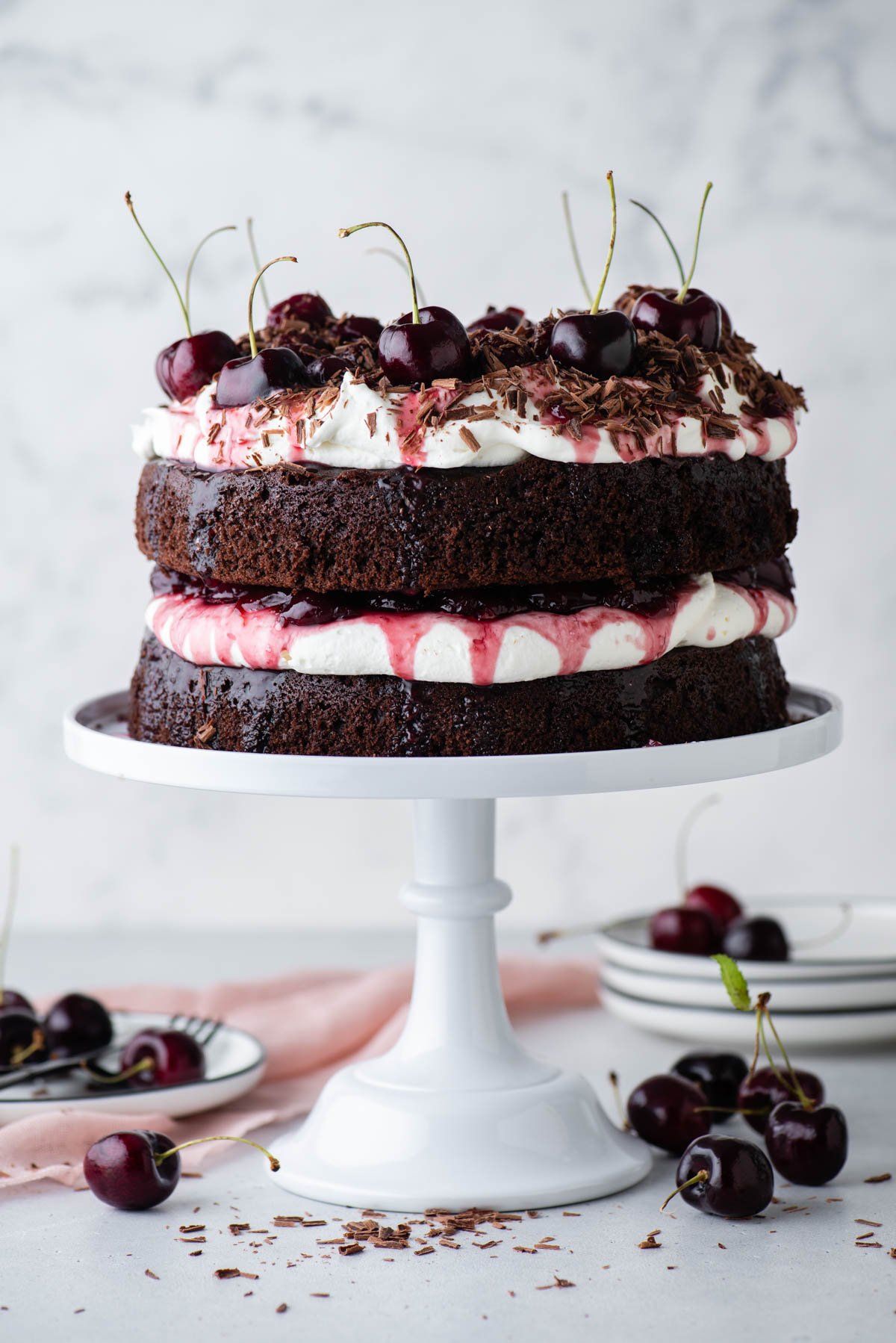 black forest cake on a white cake stand with stacks of white plates and cherries surrounding it