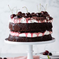 black forest cake on a white cake stand with stacks of white plates and cherries surrounding it
