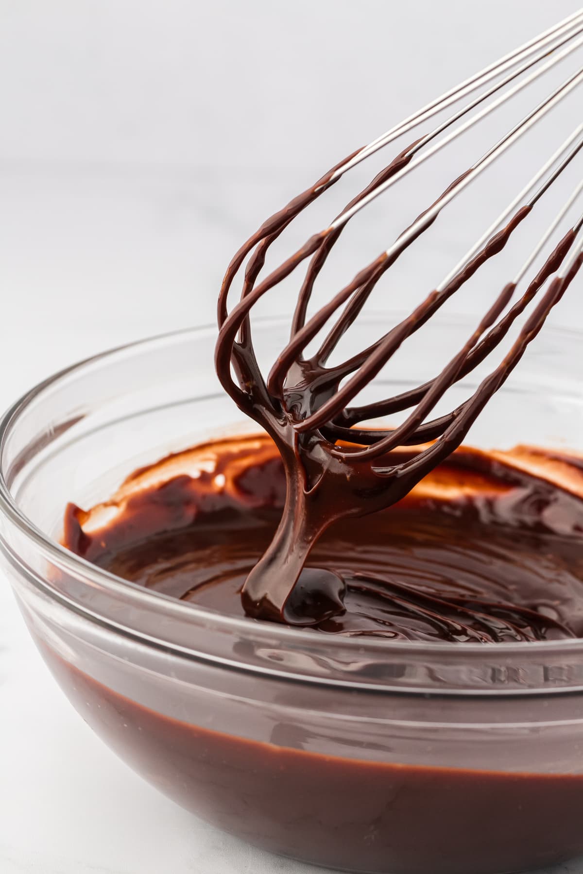 clear glass bowl full of chocolate ganache with a whisk above it drizzling ganache down into the bowl